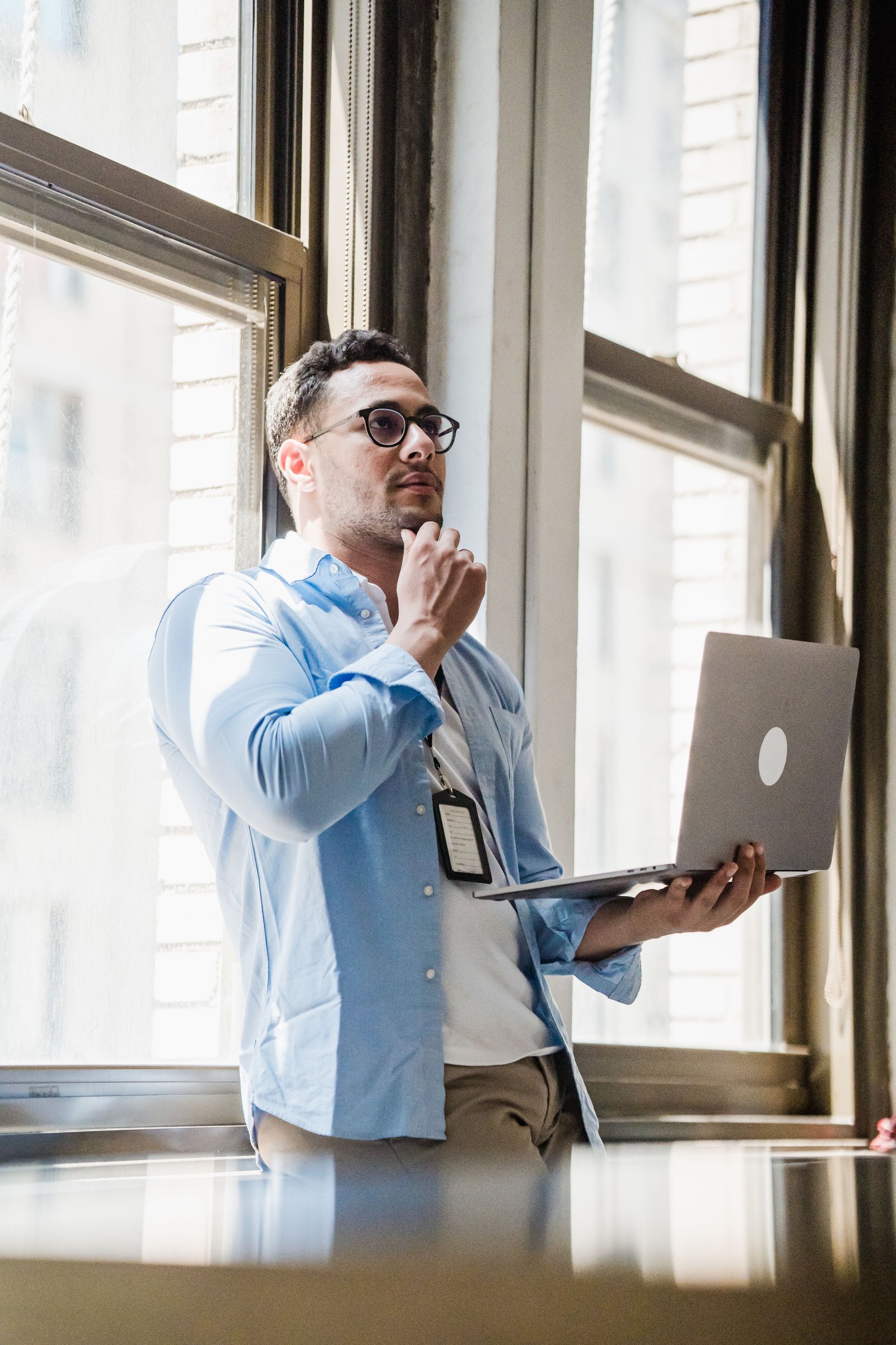 Man standing by window in office, thinking with laptop in hand.