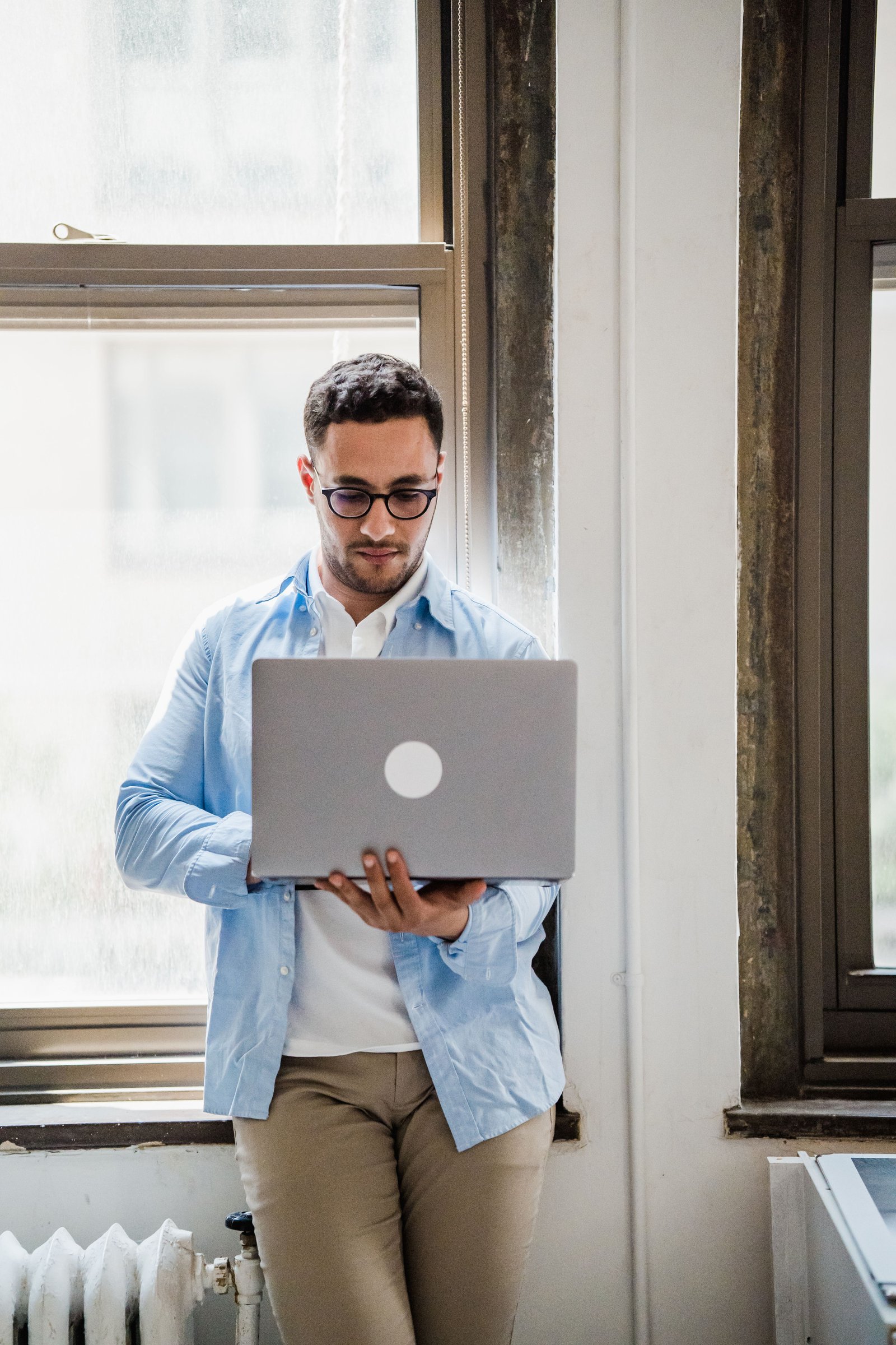 A young man in business casual attire using a laptop while leaning against an office window.