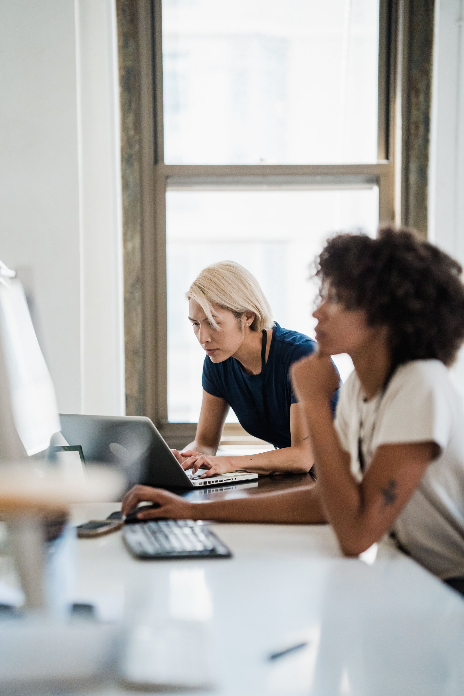 Two professional women collaborating at an office desk using laptops, focused on work.