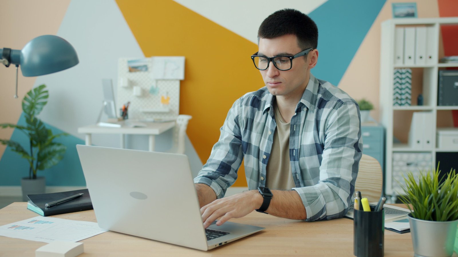 Focused young man wearing glasses, working on a laptop in a stylish office.