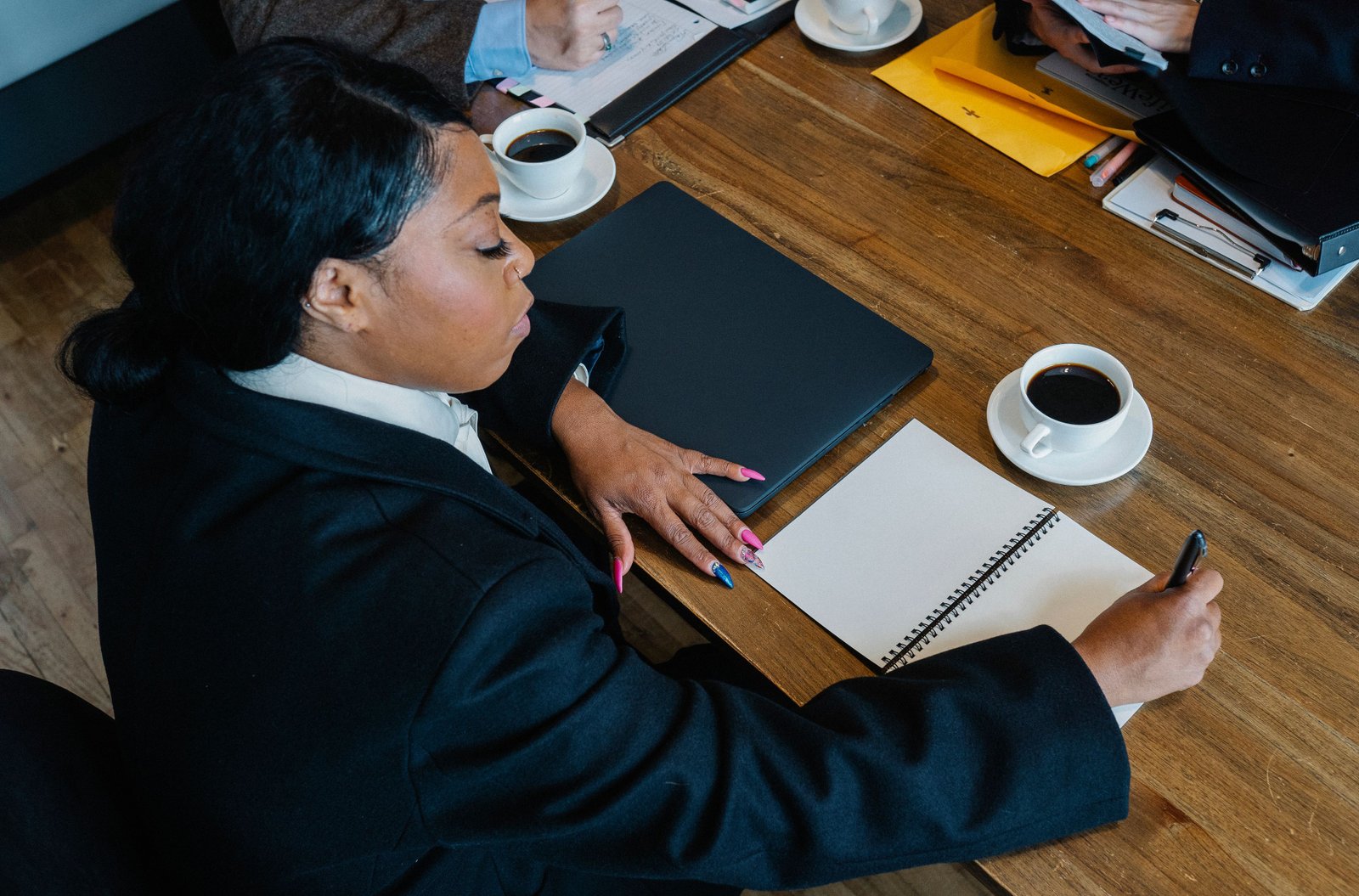 Confident businesswoman in formal attire writing notes during a meeting. Office setting with coffee and notebooks.