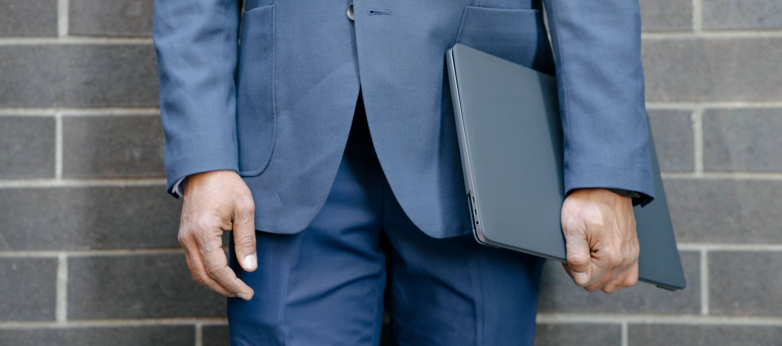 Close-up of a businessman in a blue suit holding a laptop against a brick wall.