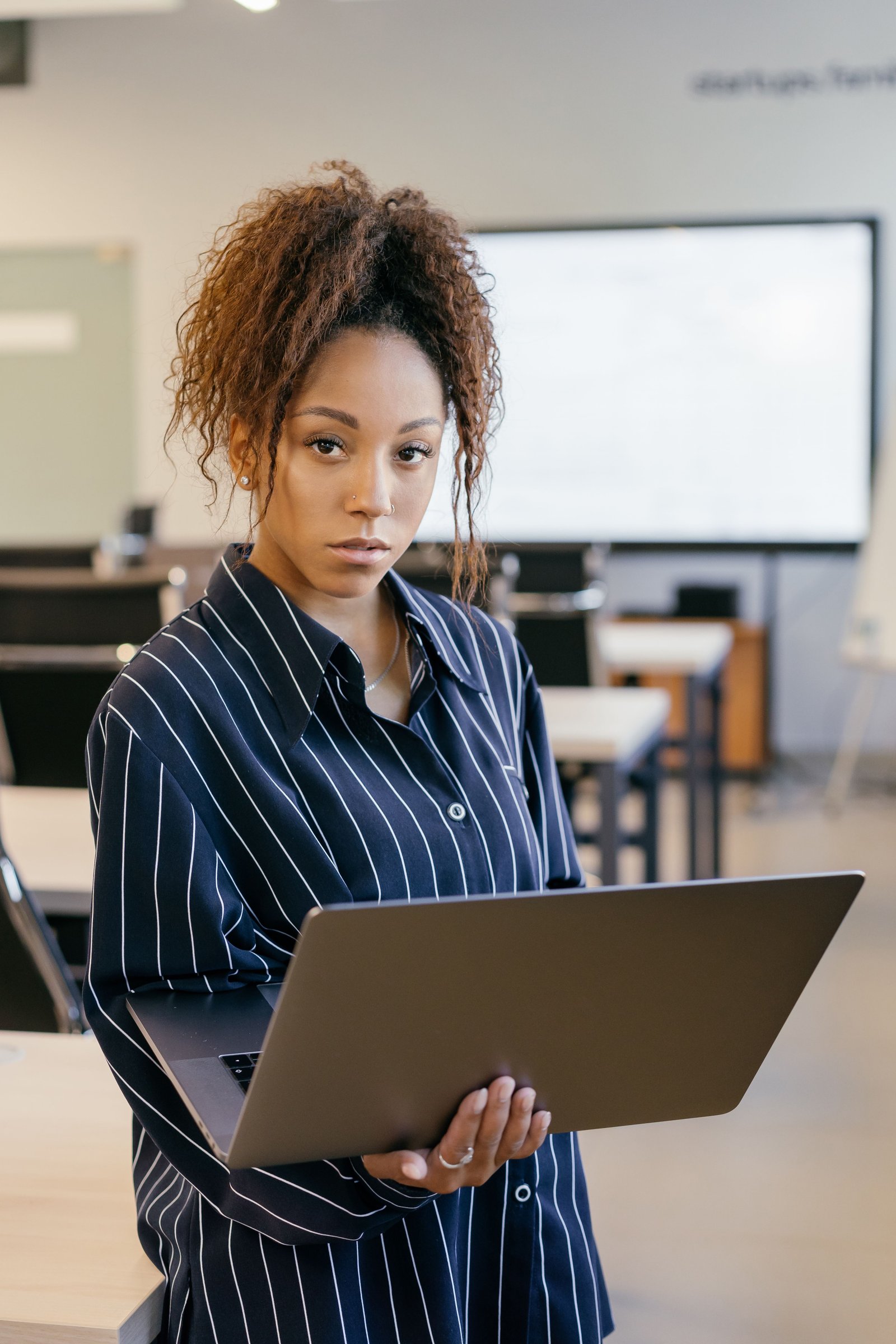 Young woman with curly hair working in an office setting, holding a laptop. Professional attire, focused expression.